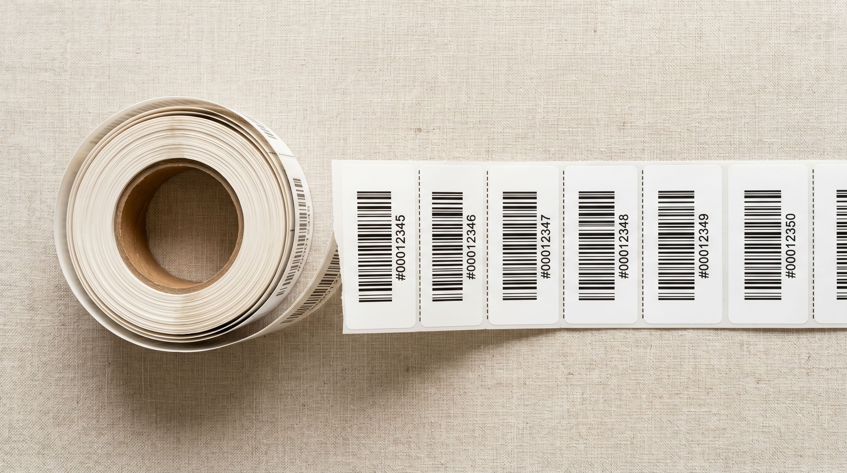 A roll of adhesive barcode sticker labels unspooled on a pale linen surface, professional studio lighting.