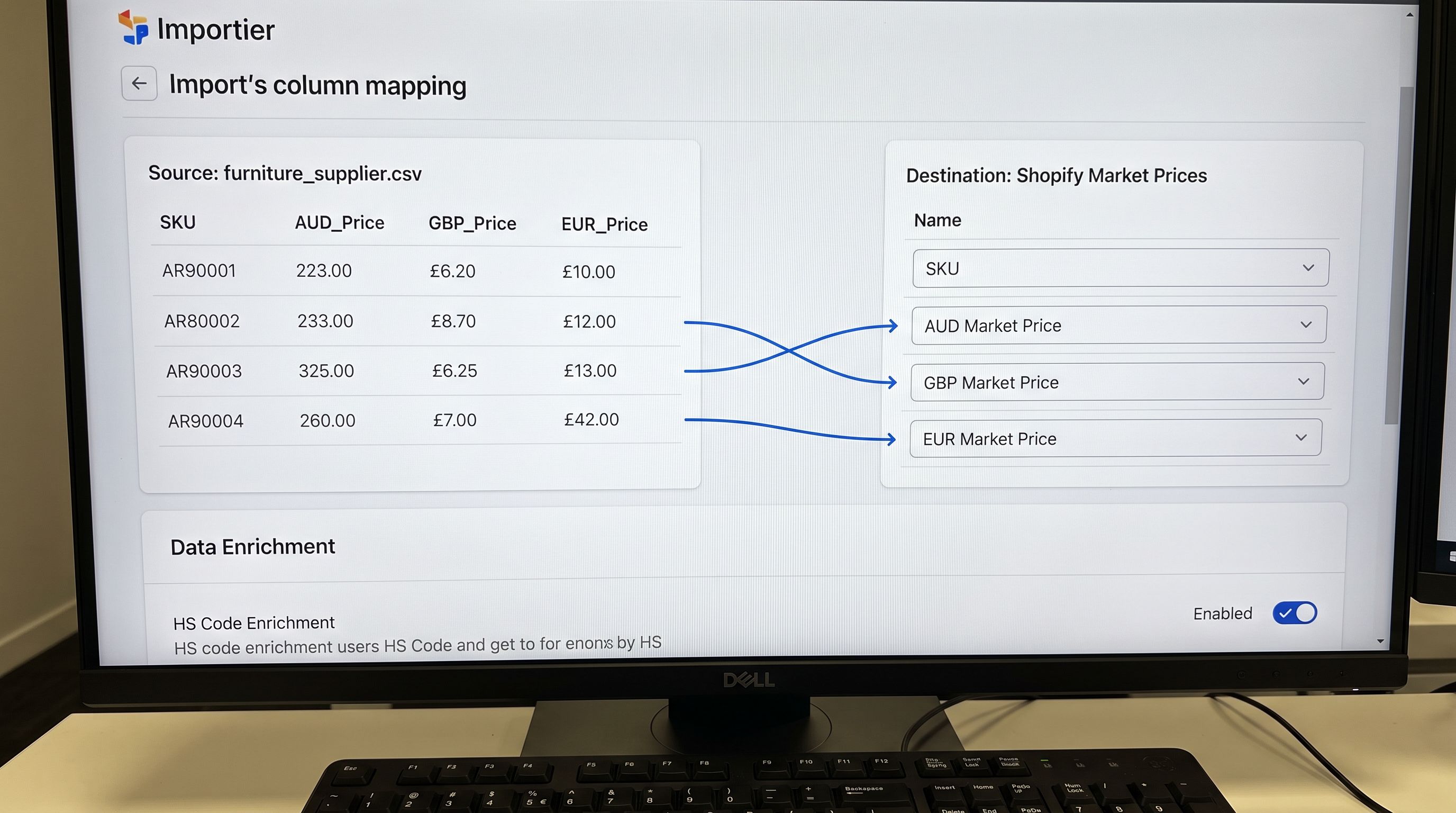 An Importier import wizard column mapping screen on a monitor showing a furniture supplier CSV with five price columns mapped to Shopify fields: price mapped to price_aud, a separate column mapped to the UK market price override, and a third mapped to the EU market price override, the Data Enrichment section below showing HS code enrichment enabled, everything in sharp focus no blur no depth of field, colour photography.