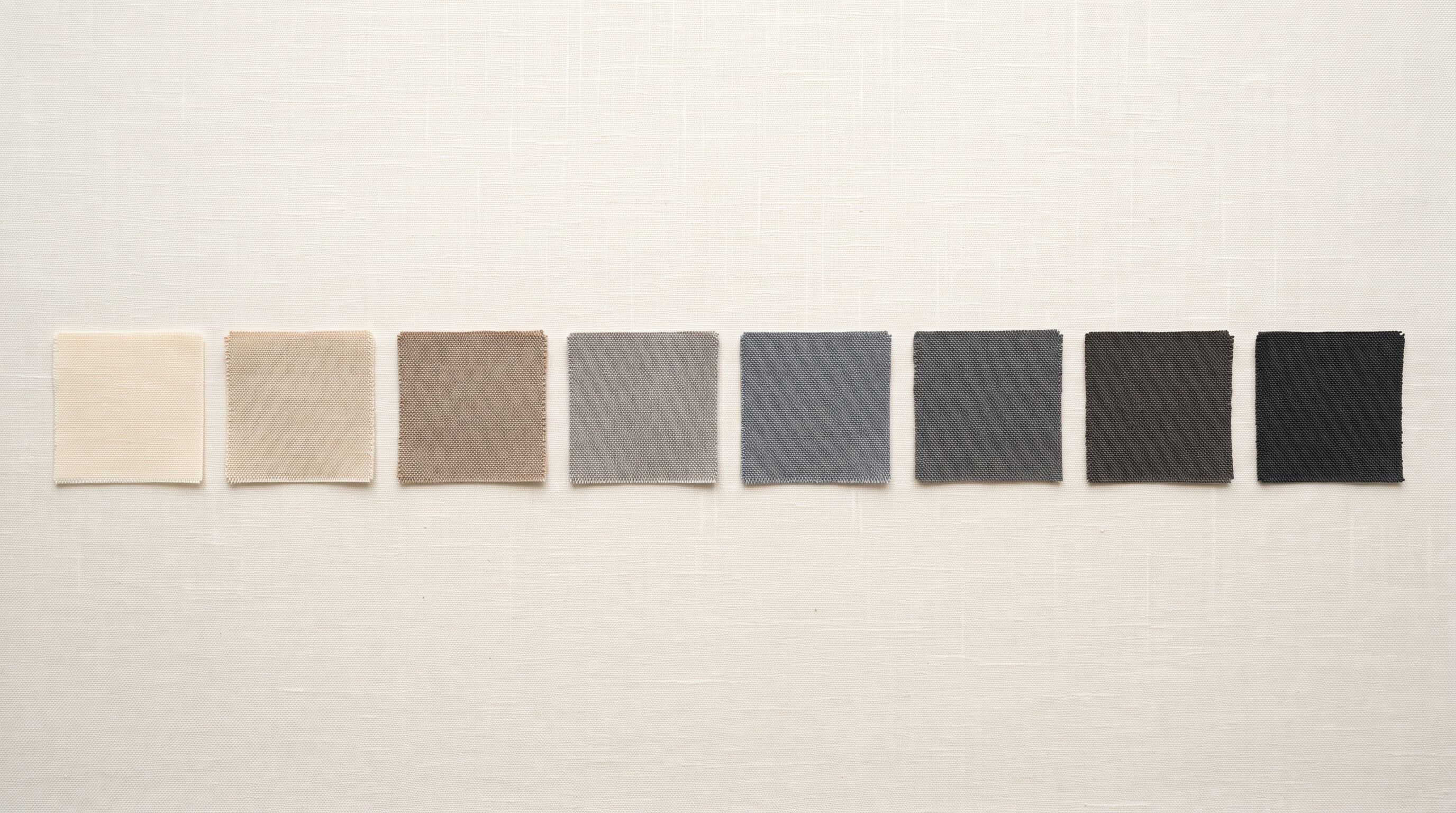 Fabric swatch samples in eight graduated colour tones from pale cream to deep charcoal on a clean linen background.