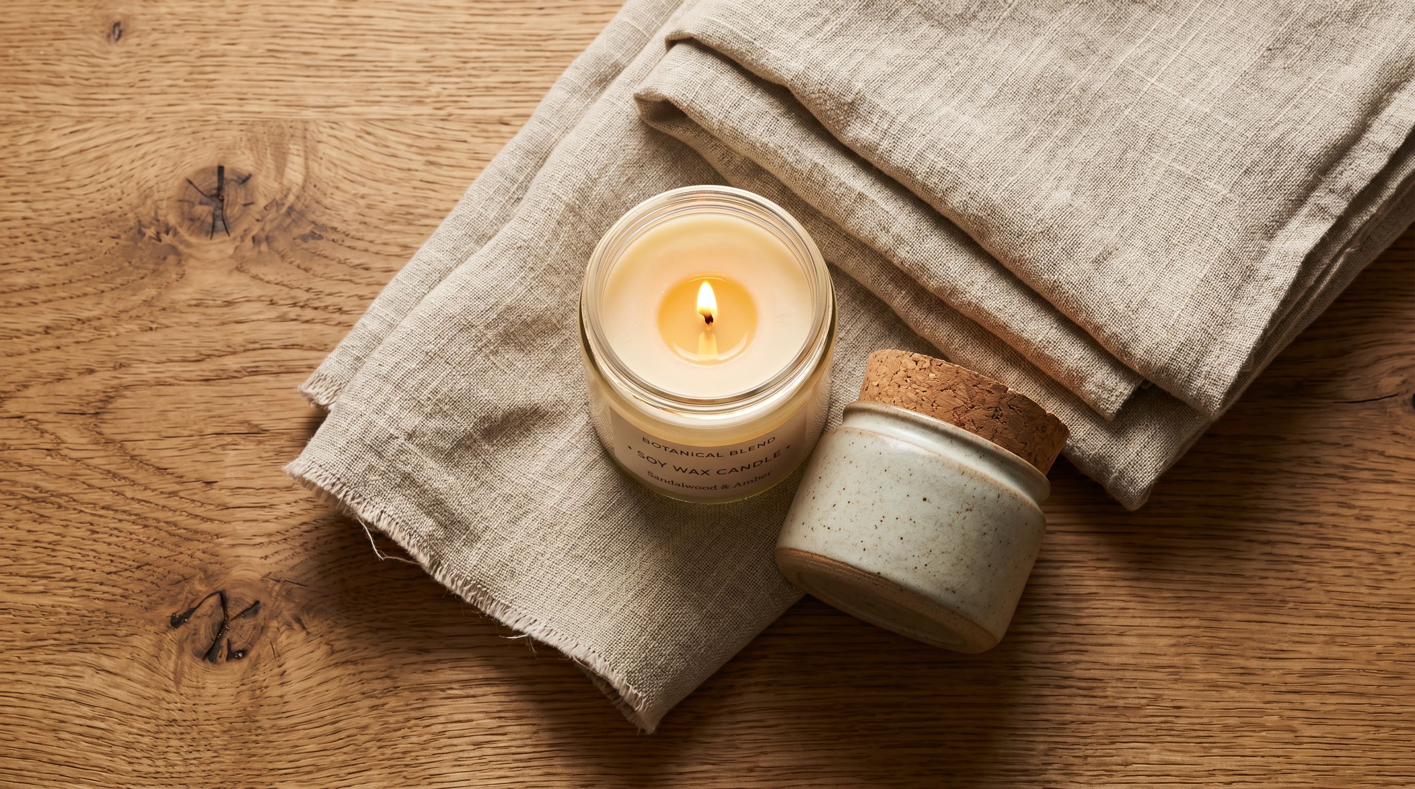 Artisan soy candle, folded linen throw, and skincare jar arranged on a natural wood surface with warm studio lighting.