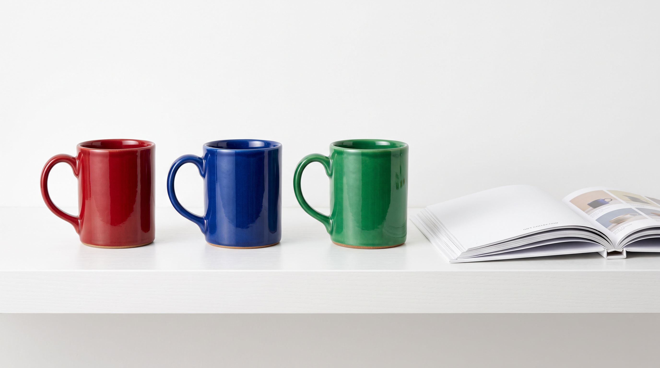 Three ceramic mugs in red, blue, and green on a white retail shelf beside a printed product catalogue document.